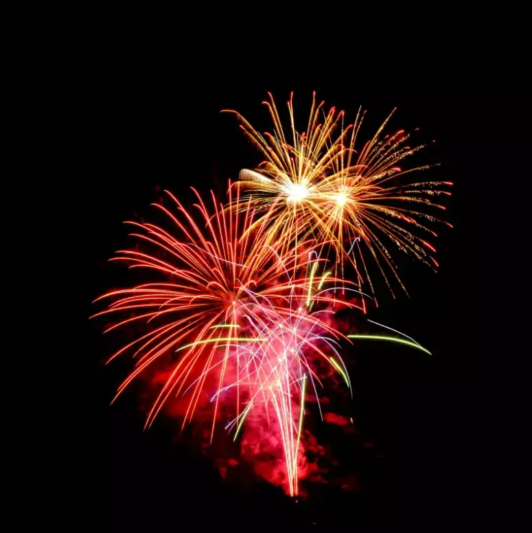 A dramatic close-up of two large fireworks bursting in the night sky. The sparks feature brilliant shades of red, orange, and yellow against a pure black background. This display represents the Firework and Night Club Shoot packages available at Kiki Beach.