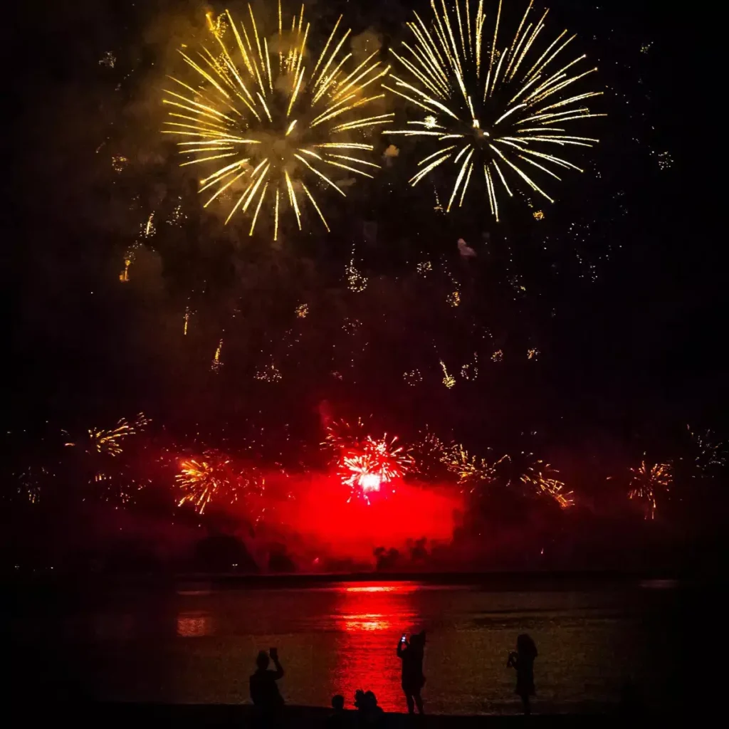 A dramatic night photograph of a group of people silhouetted on a beach, watching a massive fireworks display over the ocean. Two large golden explosions dominate the dark sky, reflecting a red glow onto the water and sand, capturing a festive moment at Kiki Beach.