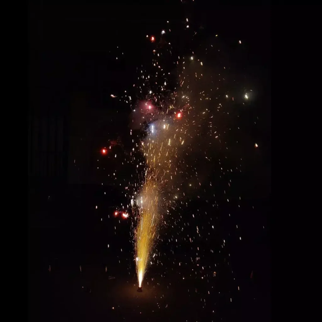 A single Roman Candle firework is actively being lit, shooting a bright, short burst of golden sparks and multiple colored pellets upwards against a dark night sky. This image represents the controlled firework entertainment available at Kiki Beach Island Resort.