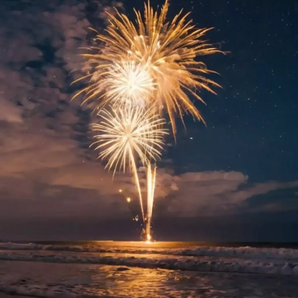 A spectacular firework display erupting from the shoreline of a beach at dusk or night. Golden sparks shoot upwards, reflecting on the wet sand and the gentle waves of the ocean. The sky above shows hints of orange and deep blue, with a few visible stars, creating a festive atmosphere at a Batam resort with beach access