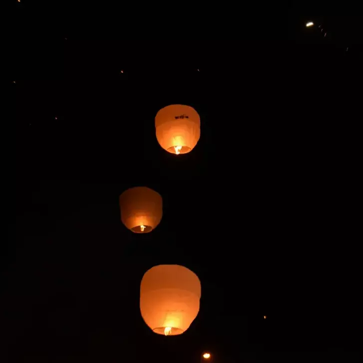 Three illuminated, orange paper wish lanterns gently ascending against a pitch-black night sky, symbolizing hope, love, and romance. The scene highlights the tranquil evening activities available at the Batam beach resort.
