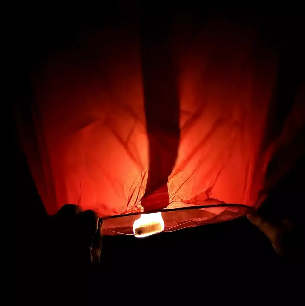 A close-up shot of a brightly glowing orange and red wish lantern being prepared for release at night. The fire source at the base is clearly visible, illuminating the paper structure from within. The dark background emphasizes the warm light of the lantern, signifying hope and romance at Kiki Beach Island Resort.