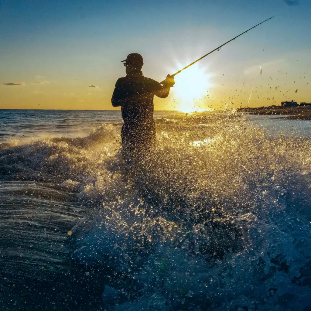 A silhouette of an angler standing in the heavy ocean surf, casting a fishing rod as the setting sun radiates a golden glow. Water splashes surround the figure. This scenic view can be associated with the fishing experience near Kiki Beach Island Resort.