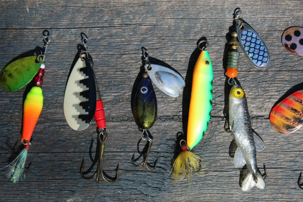 A close-up view of a selection of colorful fishing lures, spinners, and artificial bait, featuring various metal blades, plastic fish bodies, and treble hooks, all laid out on a weathered gray wooden background. This gear is typical for fishing trips near a Batam resort with beach access.