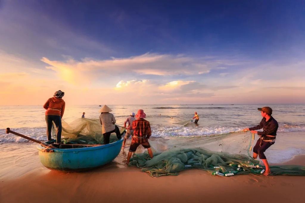 A picturesque scene of three fishermen pulling a large, heavy net onto a golden sandy shore next to a small turquoise wooden boat, likely during sunrise. The sky shows vibrant orange and blue colors. This image captures the activity often described as kiki on the beach, a common coastal sight.