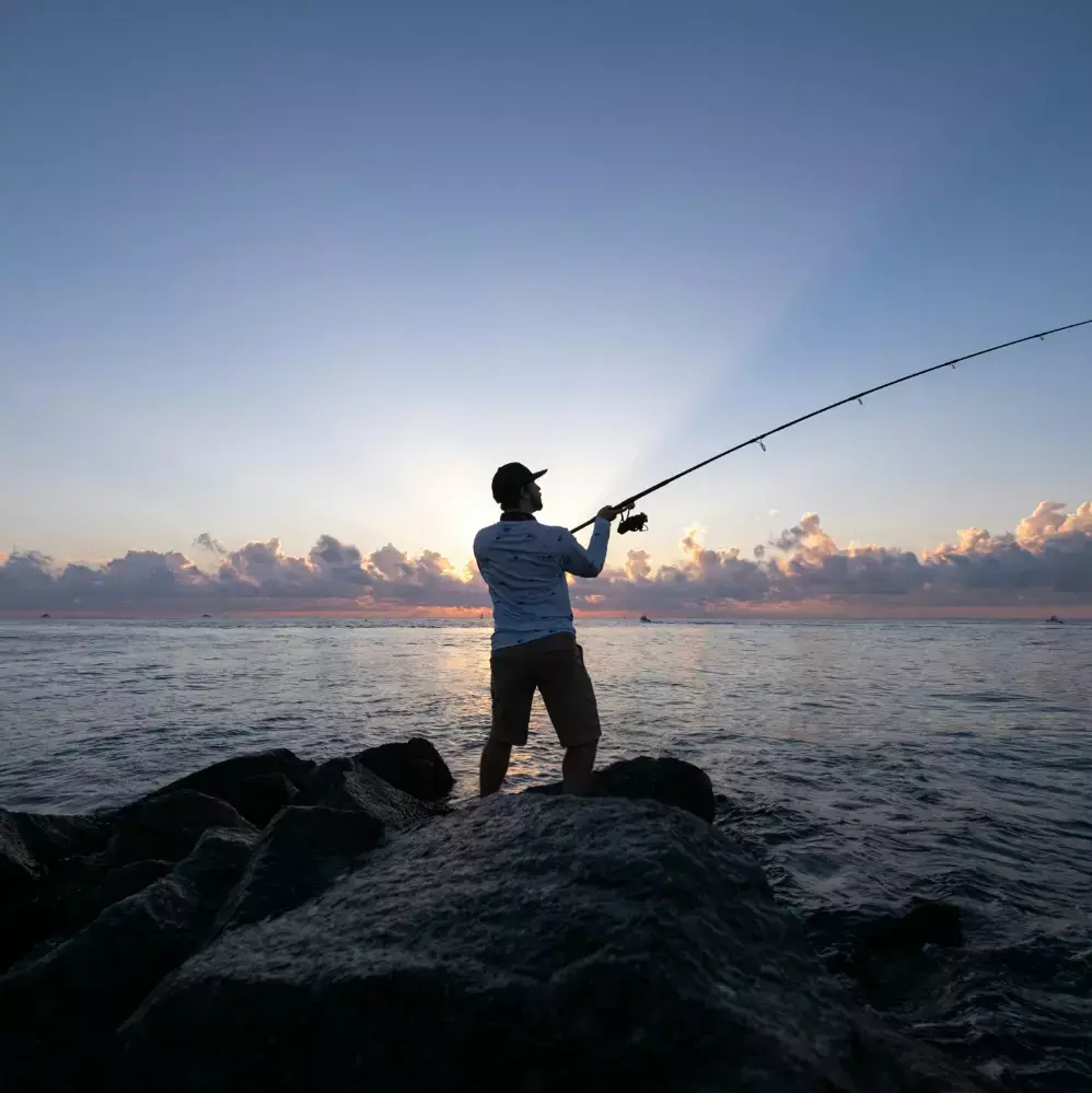 Silhouette of an individual fishing with a long rod from a rocky cliff at sunrise, with beautiful sky rays, representing an early morning fishing activity at batam beach resort.