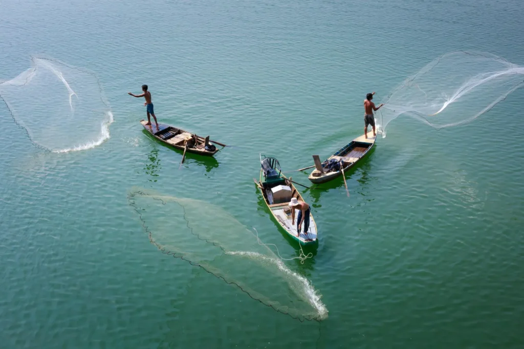 Overhead view of three local fishermen in small boats casting wide nets into the clear water, illustrating the cultural side of the batam resort with beach experience.