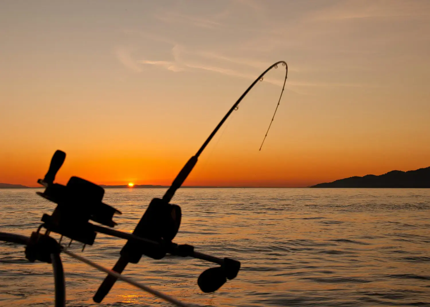 Silhouette of a fishing rod and reel against a fiery orange sunset over the water, promoting the Deep Sea Fishing activity at kiki on the beach.