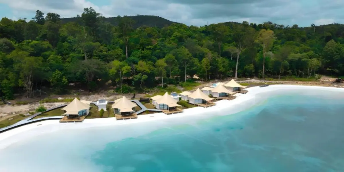 An aerial view of a secluded tropical resort featuring a row of beachfront bungalows with white thatched roofs. The resort is situated on a pristine white-sand beach, flanked by clear turquoise water and a dense, dark green mangrove forest in the background, characteristic of a Batam resort with beach access.